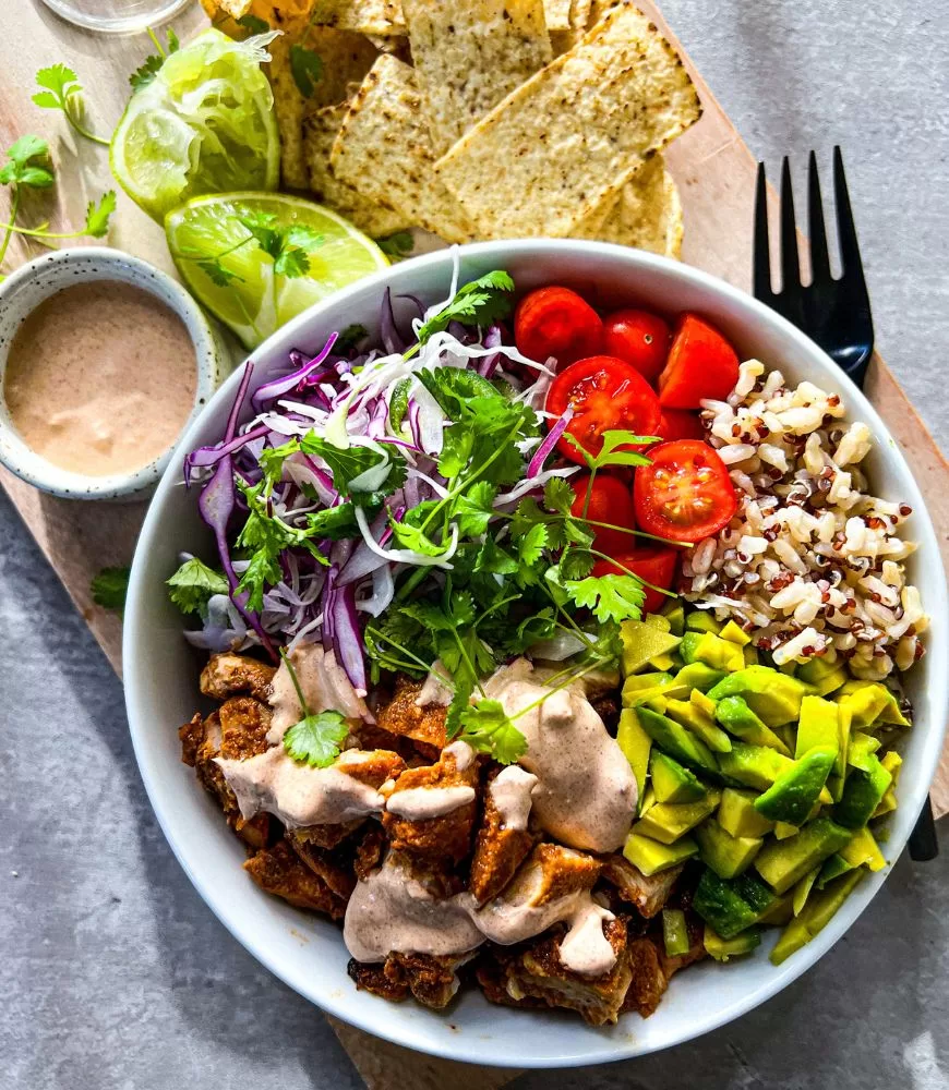 chipotle chicken and grain bowls with Mexican slaw in white bowl. Black fork, lime wedges and corn chips next to it