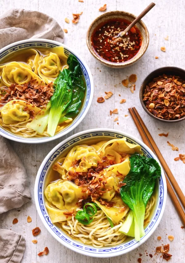 Overhead view of two bowls of wonton noodle soup with egg noodles, wontons and bok choy in clear broth, styled on a white timber surface with a neutral cloth, wooden chopsticks, chili oil and crispy shallots scattered for a slightly messy, rustic look in bright natural light.