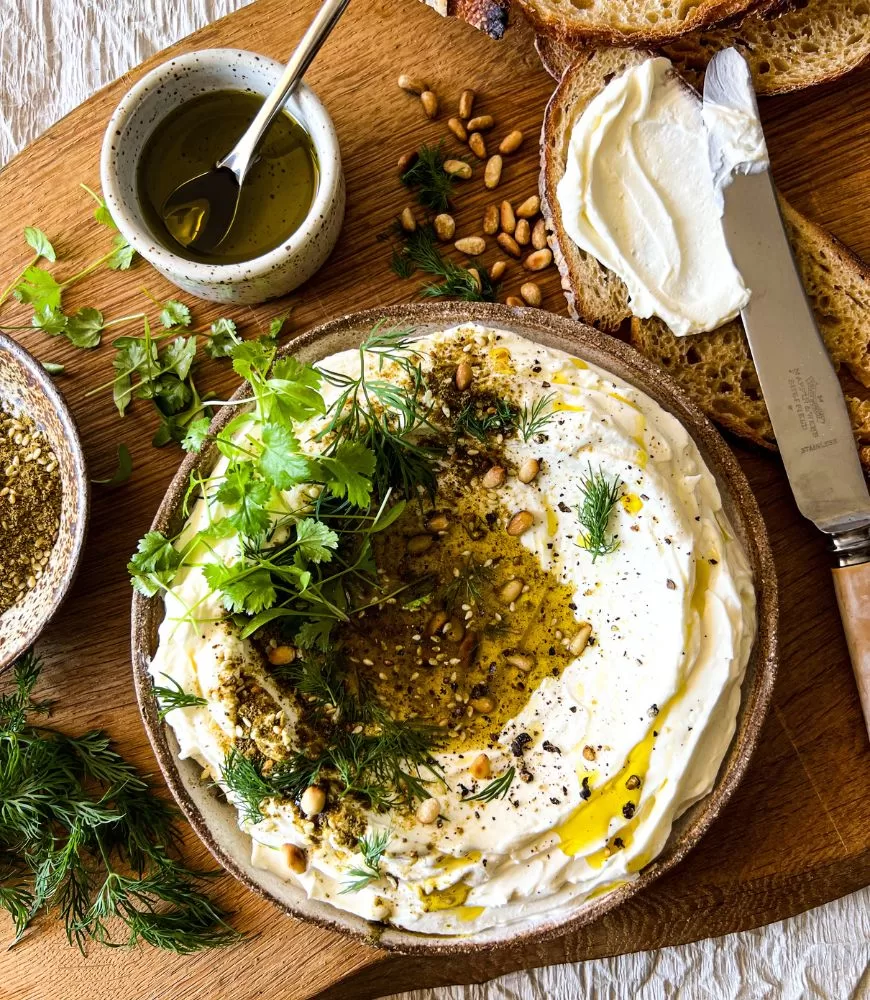 labneh dip in bowl  with herbs on wooden board with sourdough bread slices, knife, herbs, pine nuts, olive oil and zatar around it