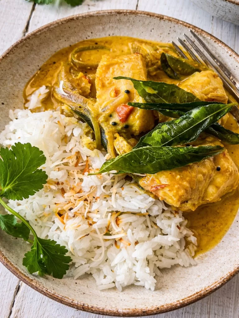 Close-up of Kerala fish curry in coconut sauce served with basmati rice in a ceramic bowl, garnished with curry leaves and fresh herbs on a white timber table in natural light.