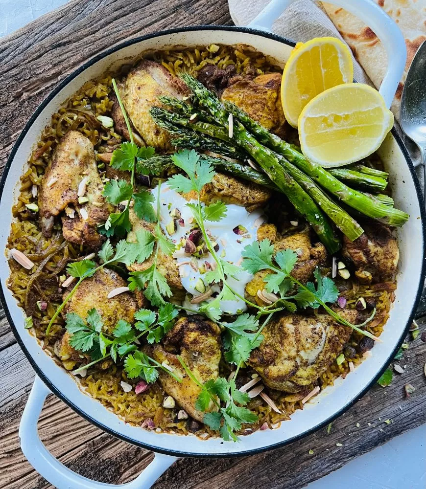 white baking dish filled with Middle Eastern chicken and rice topped with yoghurt, nuts, asparagus and lemon on wooden background