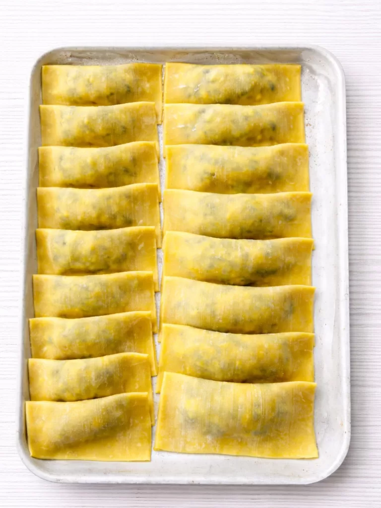 Tray of uncooked spinach and ricotta ravioli arranged in rows on a flour-dusted baking sheet over a white timber background.