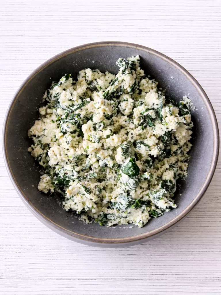 Grey bowl filled with spinach and ricotta filling on a white timber background, showing the creamy texture and flecks of green spinach.