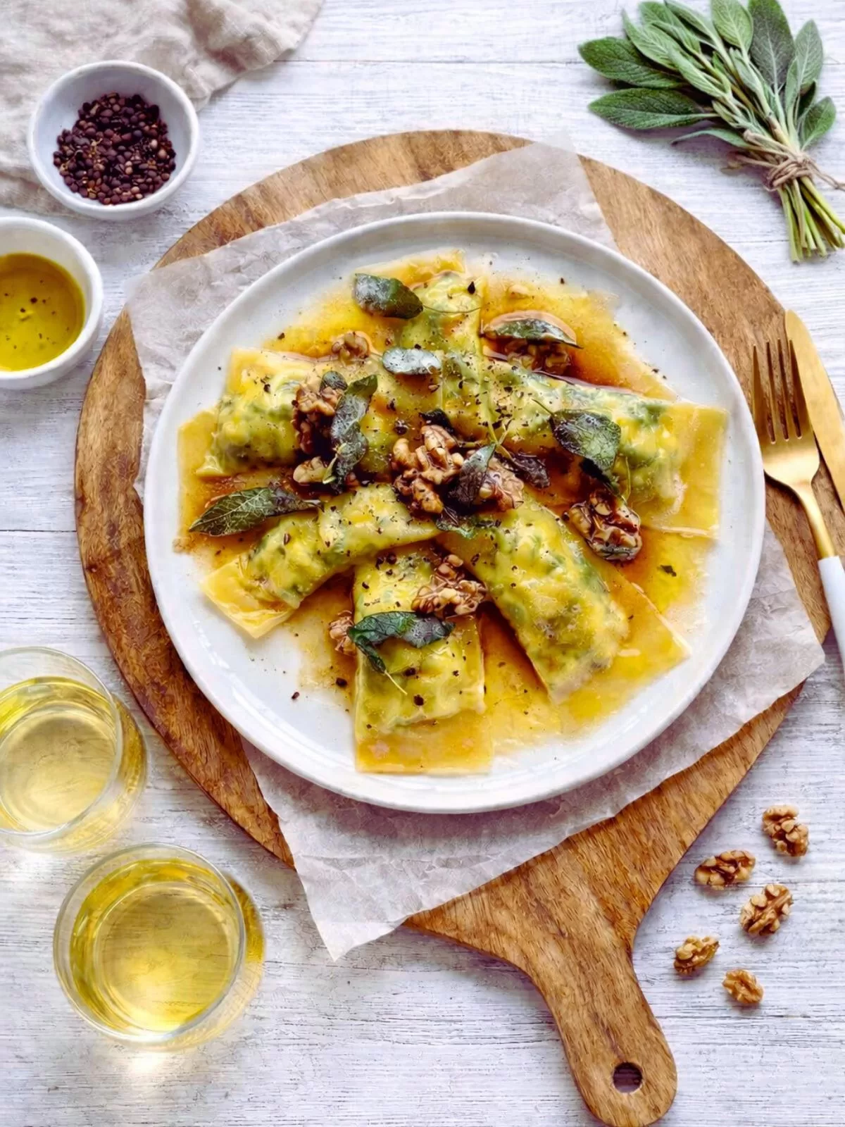 Spinach and ricotta ravioli on a white plate with butter sauce, crispy sage and walnuts, set on a wooden board with glasses of white wine and fresh sage on a white timber table.