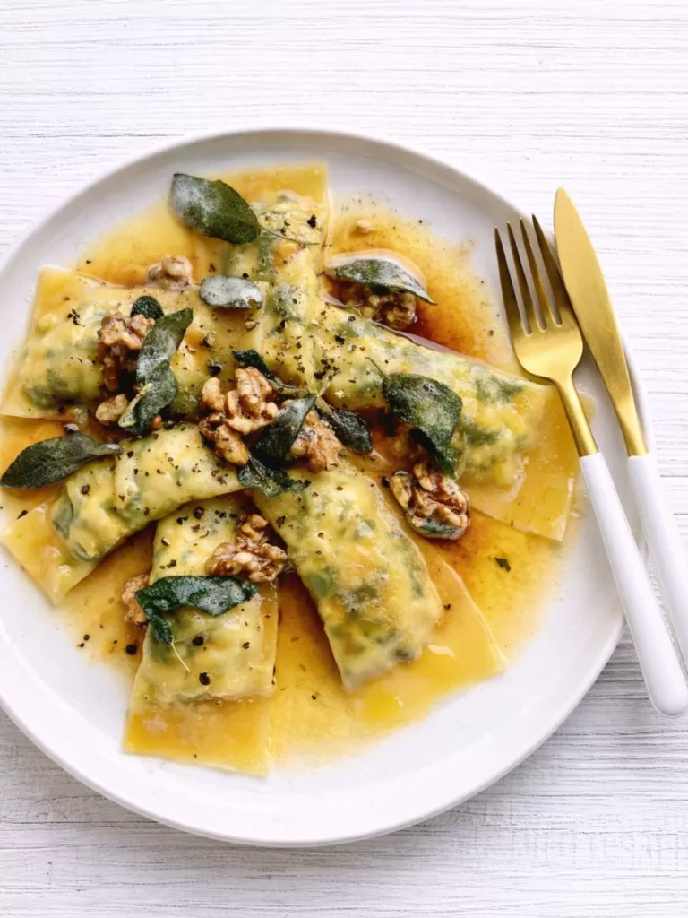 Spinach and ricotta ravioli in butter sauce with crispy sage and walnuts on a white plate, set on a white timber background with a white and gold knife and fork on the right.