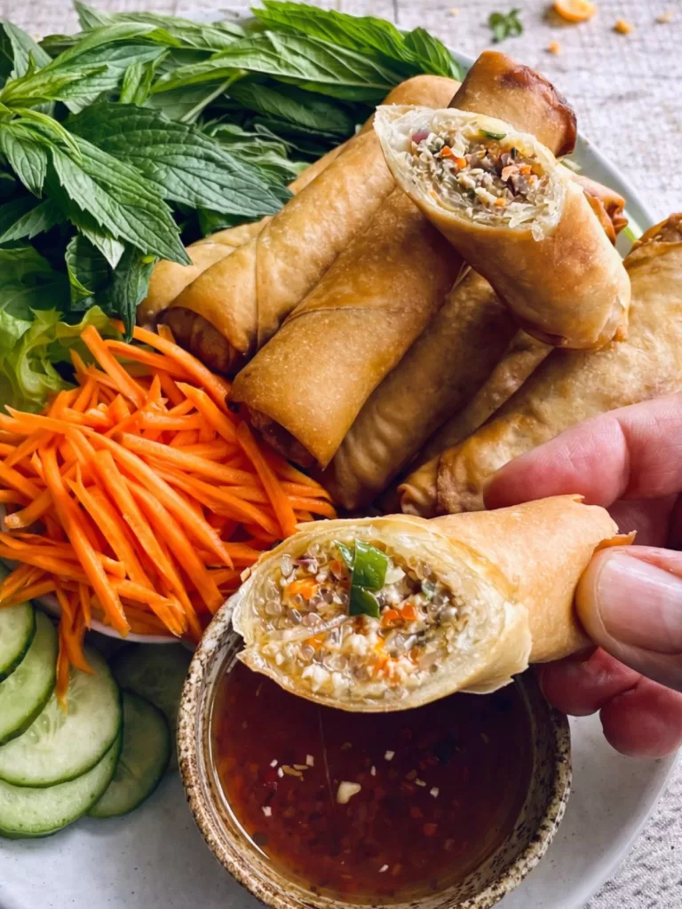 Close-up of Vietnamese spring rolls on a neutral plate over a white timber background, with one roll held and dipped into sweet chilli sauce, showing a filling of glass noodles, carrot and herbs in bright natural light