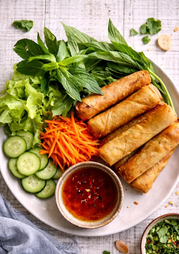 Overhead view of crispy Vietnamese spring rolls on a neutral off-white plate, served with fresh mint, lettuce, cucumber and shredded carrot, plus a bowl of sweet chilli dipping sauce on a white timber background in natural light.