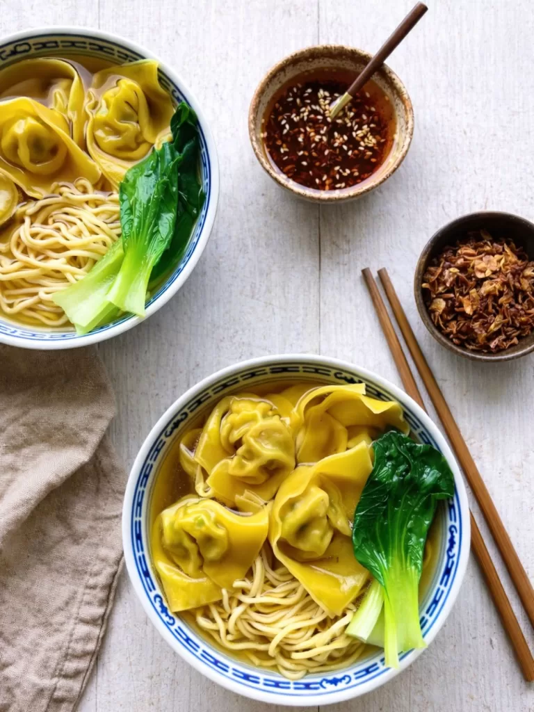 Overhead view of two bowls of wonton noodle soup with egg noodles, plump wontons and bok choy in clear broth, styled on a white timber surface with a neutral linen napkin, wooden chopsticks and small bowls of chilli oil and crispy shallots in bright natural light.