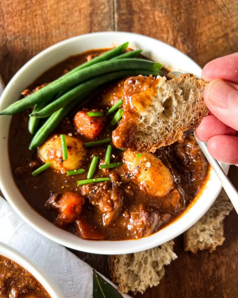 Beef in red wine in a bowl with a hand dunking bread into it