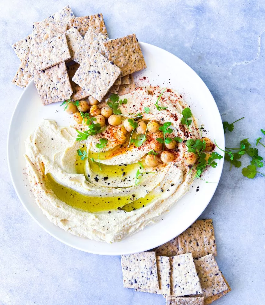 white plate with hummus swirled on it with chickpeas, paprika, pepper and garnish. Biscuits around it