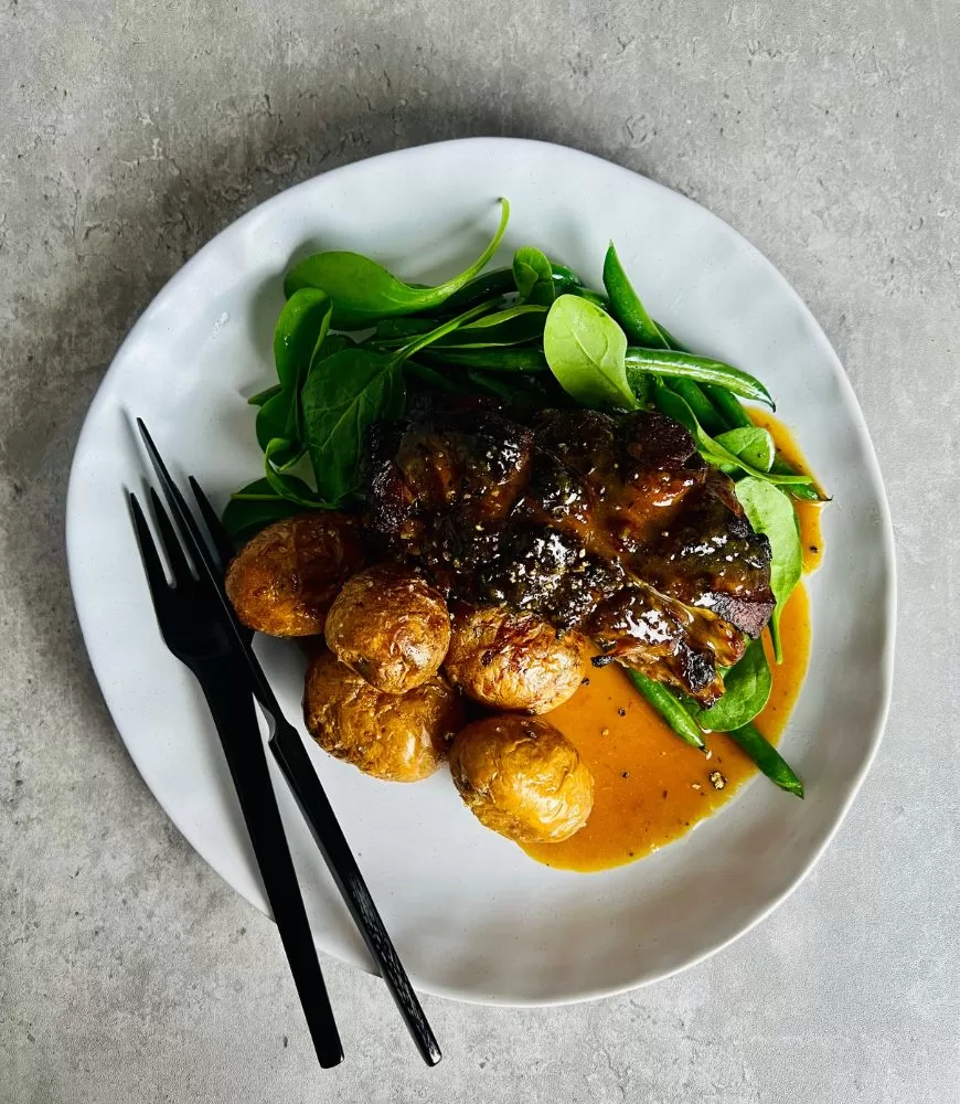 lamb shoulder on white plate with potatoes, beans and spinach with gravy. Black knife and fork. Grey concrete background