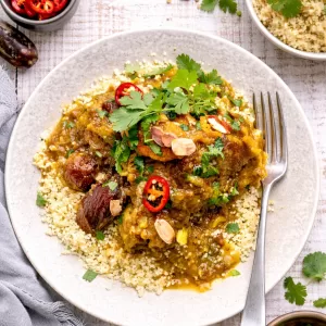 Overhead view of Moroccan chicken tagine with dates and carrots served over couscous on a white timber background, styled with small bowls of coriander and sliced chilli, scattered herbs and a fork in natural light.