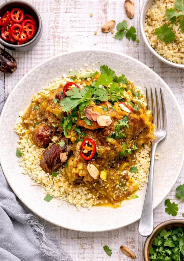 Overhead view of Moroccan chicken tagine with dates and carrots served over couscous on a white timber background, styled with small bowls of coriander and sliced chilli, scattered herbs and a fork in natural light.