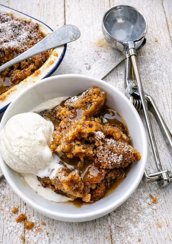 Sticky date pudding served in a white bowl with caramel sauce and a scoop of vanilla ice cream, dusted with icing sugar, on a white timber background with a baking dish and ice cream scoop nearby in natural light.