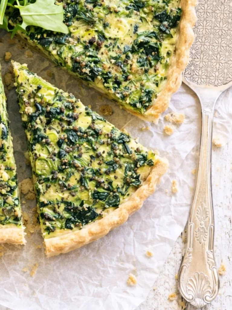 Close-up of sliced spinach and feta quiche on parchment paper over a white timber table, with a flaky golden crust, green herb-filled egg mixture, scattered crumbs, and a vintage silver pie server alongside.