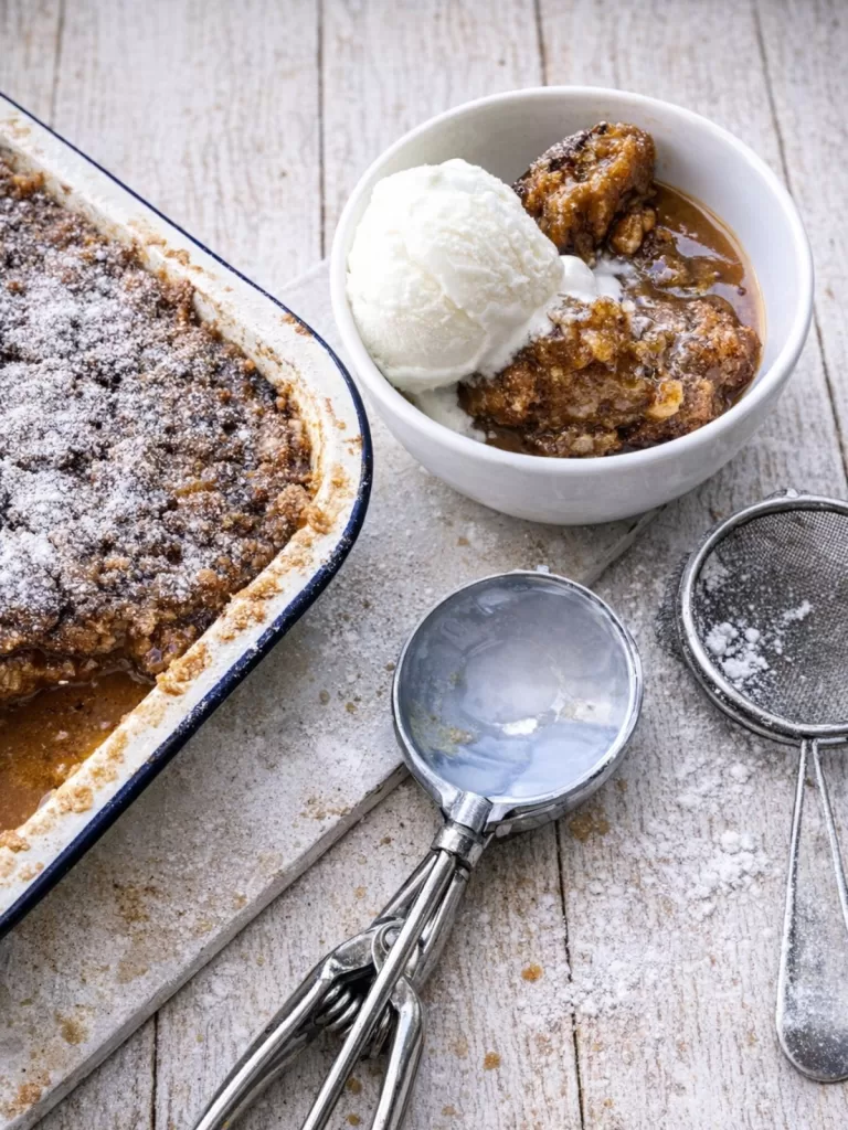 sticky date pudding in baking dish, bowl of pudding with ice cream and ice cream scoop in fore ground. white timber background.