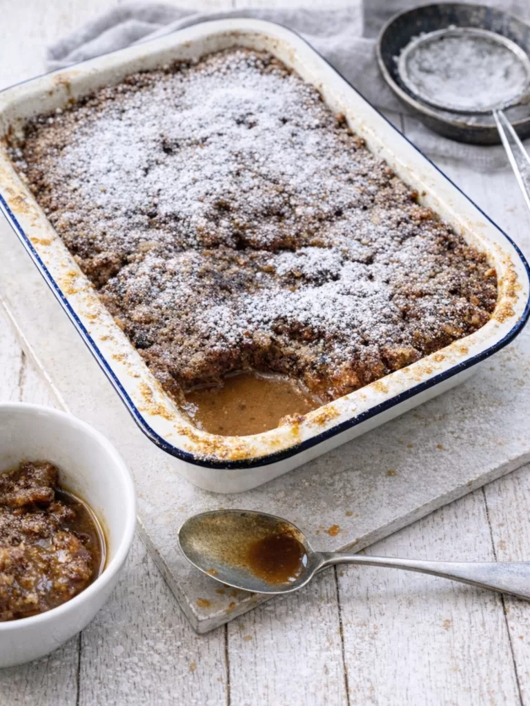 Self-saucing sticky date pudding in a white enamel baking dish, dusted with icing sugar, with caramel sauce visible, on a white timber table and pale board in natural light.