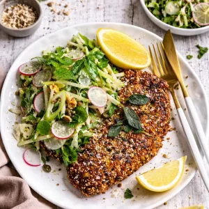 Crispy chicken schnitzel coated in panko breadcrumbs and mixed seeds on a white plate over a white timber background, served with fresh herb salad and pale lemon wedges in natural light with scattered crumbs.