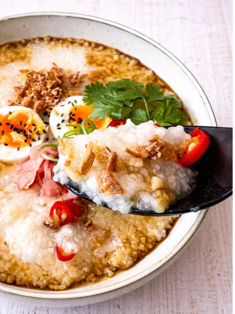 Close-up of a spoonful of creamy congee with crispy shallots and chilli above a bowl of congee topped with jammy eggs and fresh coriander on a white timber surface.