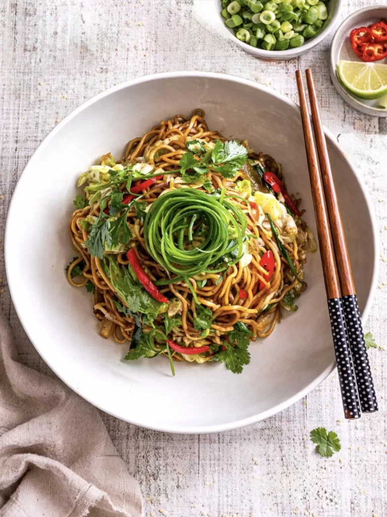 Overhead view of soy sauce noodles in a white bowl on a white timber table, topped with spring onion curls, coriander and red chilli, with chopsticks and small bowls of soy sauce and chopped spring onions, scattered sesame seeds and herbs in natural light.