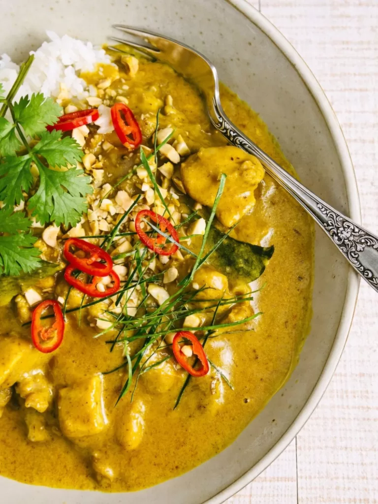 cream satay chicken curry in a beige bowl with fork on white timber background. Garnished with kaffir lime leaves, red chilli and coriander.