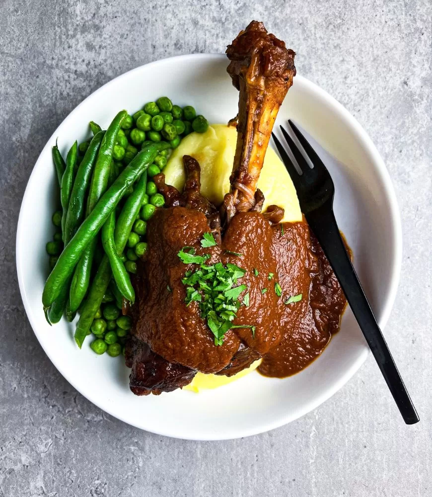 lamb shank in red wine with mash, peas and beans in white bowl grey background