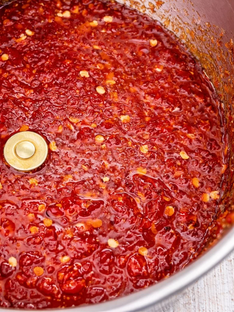 Close-up of chunky homemade chilli jam simmering in a pot, with visible chilli seeds and a glossy red texture