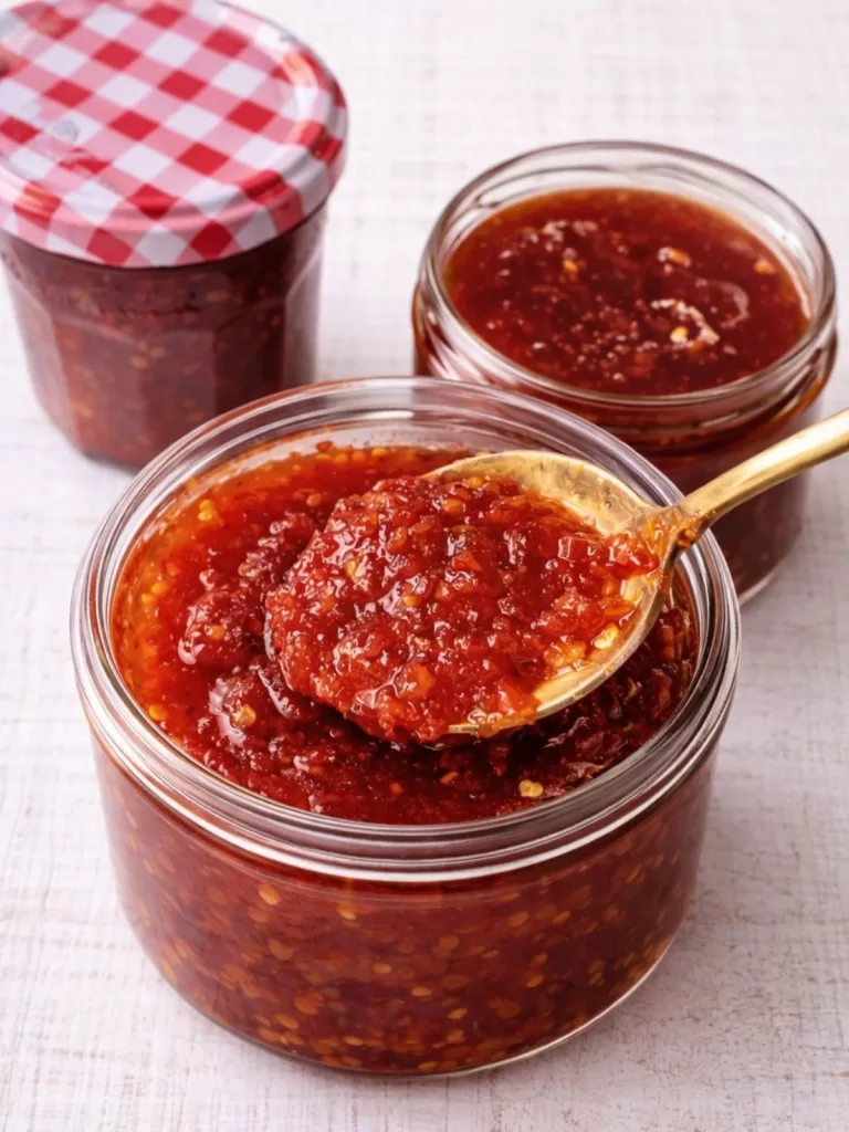 Open jars of glossy red chilli jam with visible chilli seeds, one topped with a gold spoonful, on a white timber background.