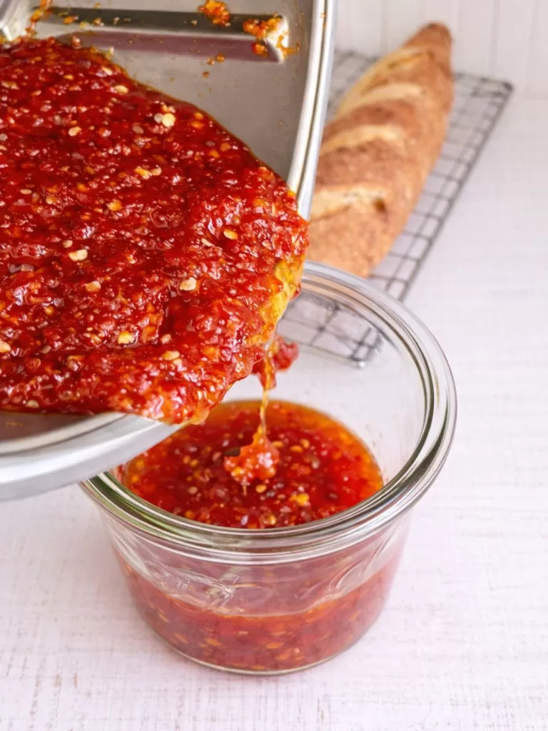 Thick homemade chilli jam being poured from a saucepan into a glass jar, with visible chilli seeds, on a white timber background.
