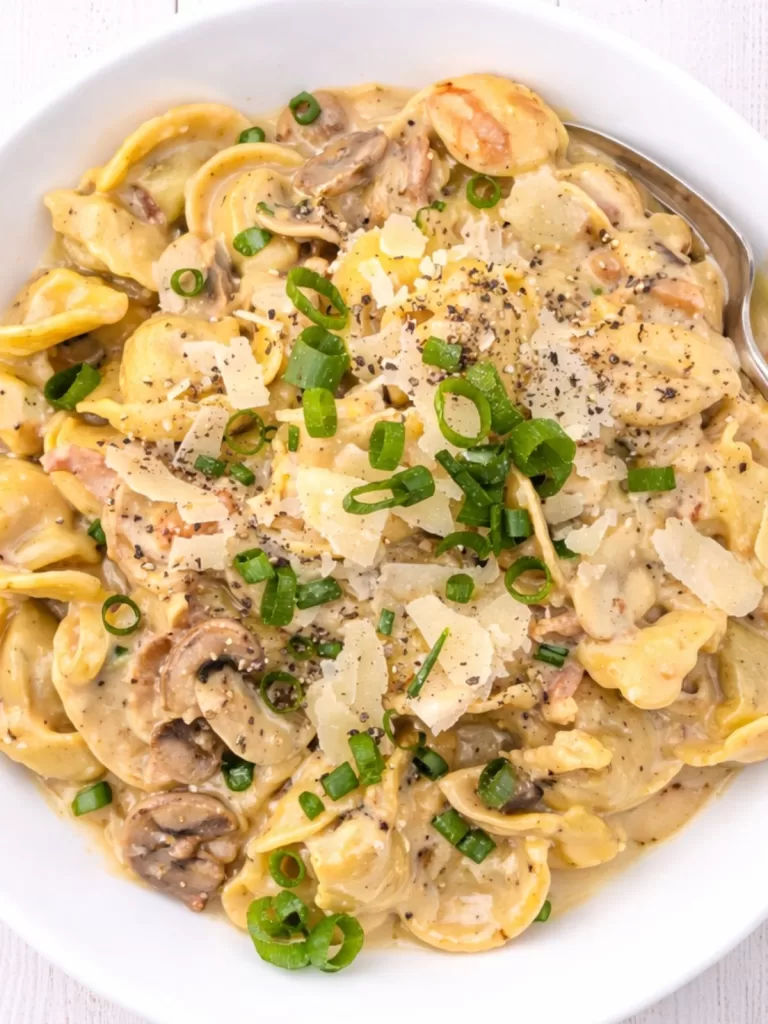 Overhead view of creamy French onion tortellini with bacon and mushrooms in a white bowl, topped with shaved Parmesan, black pepper and sliced green onions, on a bright white timber surface in natural light.