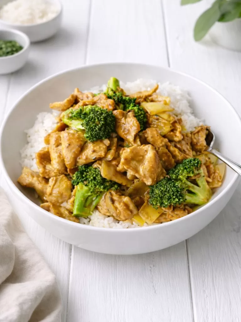 Satay chicken stir fry served over white rice in a white bowl, topped with broccoli and onions, set on a white timber background with soft natural light and minimal styling.