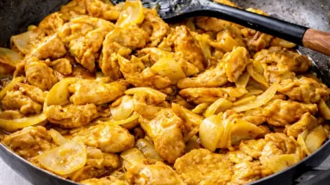 Black wok filled with golden curry chicken and onions on a white timber table, with a teaspoon of curry powder, fresh coriander, a neutral linen napkin, and a small bowl of sliced spring onions nearby.