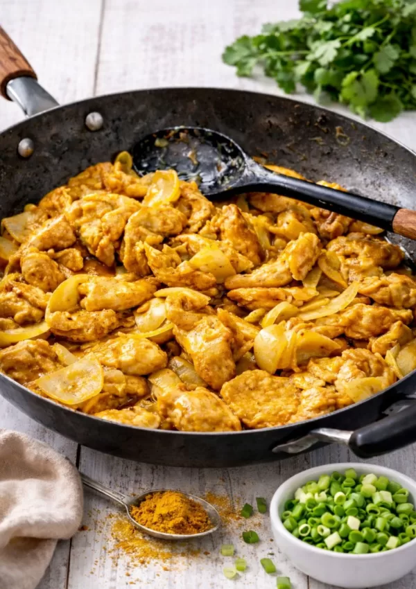 Black wok filled with golden curry chicken and onions on a white timber table, with a teaspoon of curry powder, fresh coriander, a neutral linen napkin, and a small bowl of sliced spring onions nearby.
