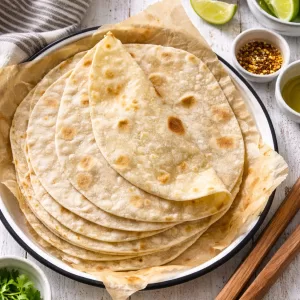 Stack of homemade flatbreads on parchment paper in a white enamel tray, styled on a white timber table with a striped tea towel, wooden tongs and small bowls of salt, olive oil and lime wedges.