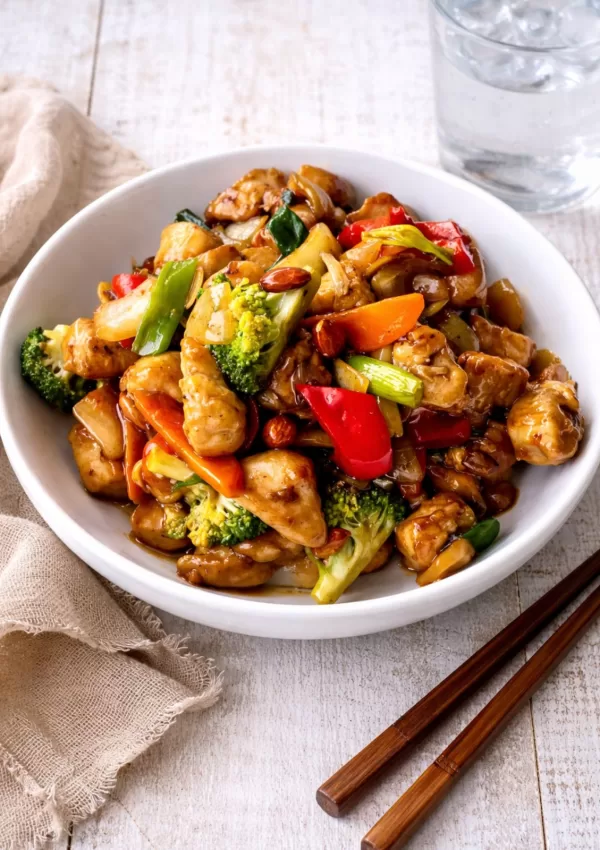 Chinese chicken with roasted almonds in a glossy savoury sauce, served with vegetables in a bowl on a white timber surface, with chopsticks, a napkin and a glass of water.