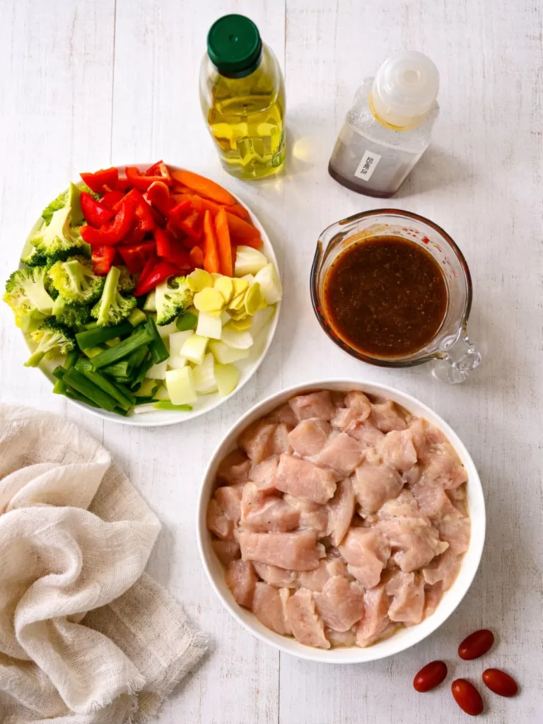 “Overhead view of Chinese chicken stir-fry ingredients on a white timber surface, including diced raw chicken, chopped broccoli, capsicum, carrots, spring onions, garlic, oil and a bowl of sauce