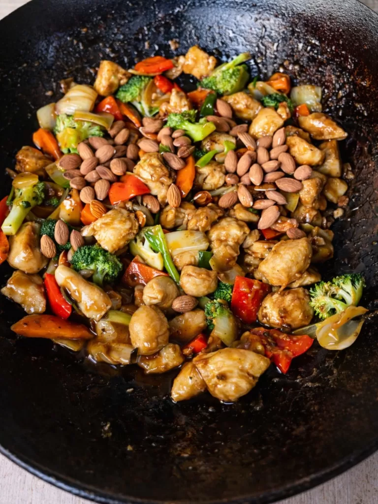 Chinese chicken stir-fry with broccoli, capsicum, carrots and whole almonds in a glossy brown sauce, cooking in a black wok.