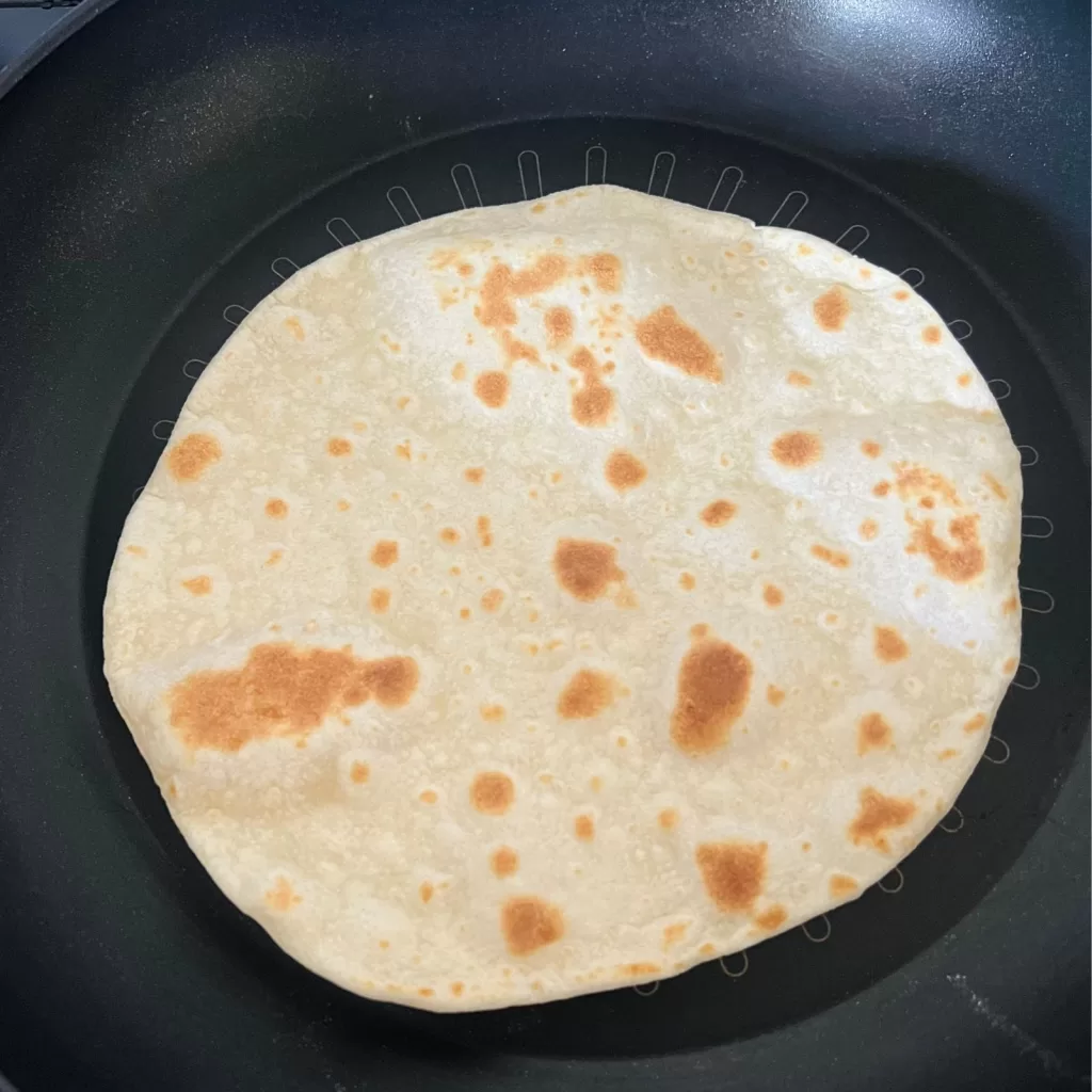 A flatbread cooking in a frying pan with golden brown spots forming on the surface.