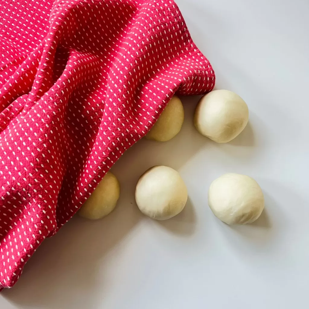 Small dough balls resting on the bench under a red tea towel, ready to be rolled out.