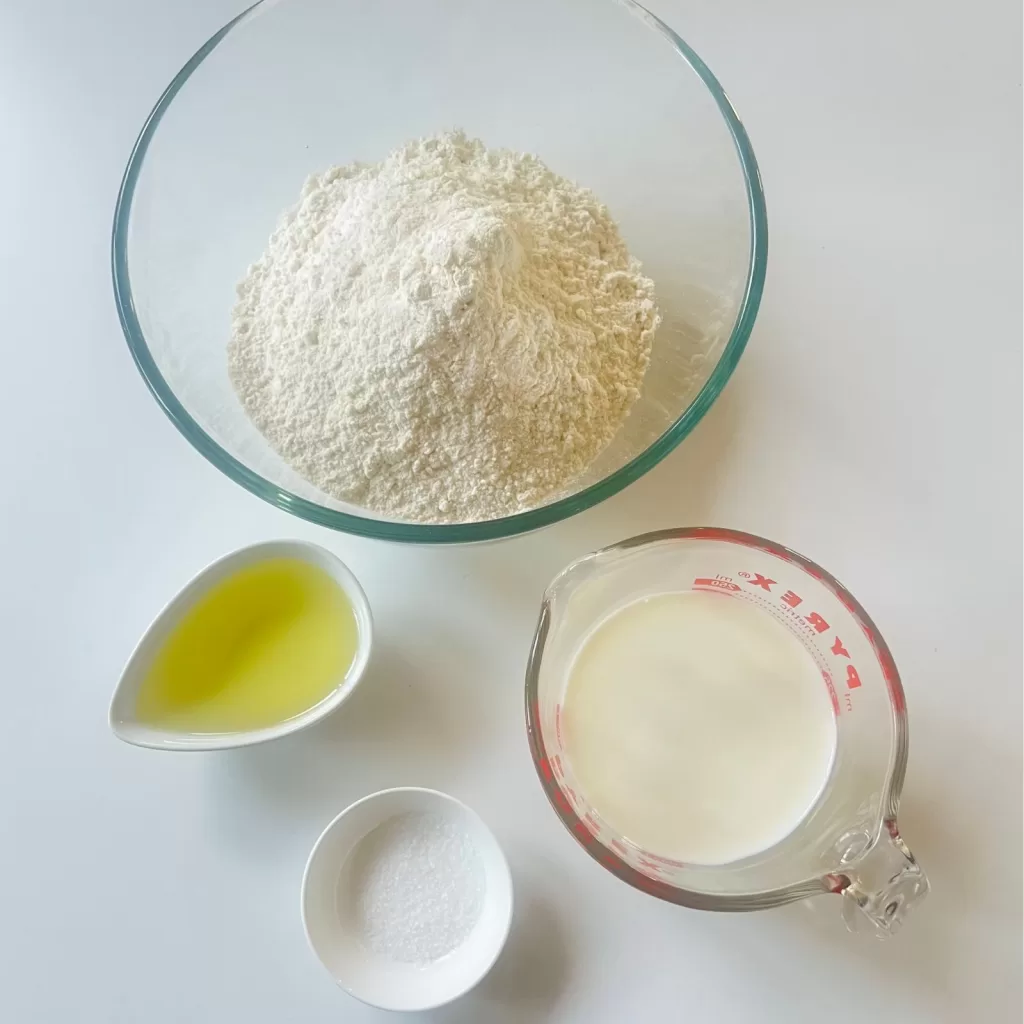 Flour, olive oil, salt and a jug of milk arranged on a white bench ready for making flatbread dough.