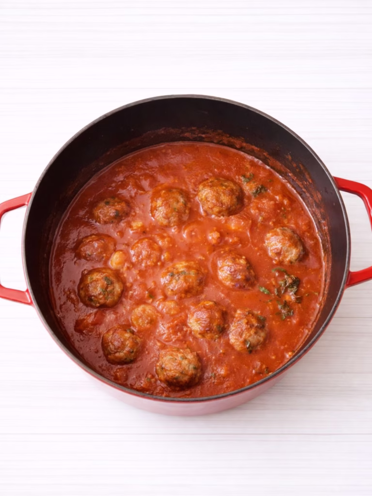Meatballs simmering in rich tomato sauce in a red pot on a white timber background