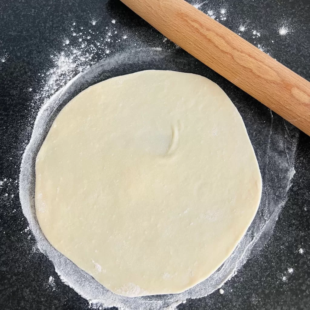 A flatbread dough round being rolled out on a floured bench with a rolling pin nearby.