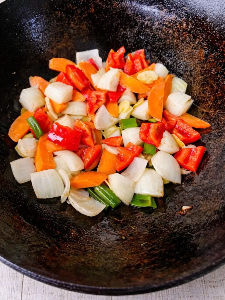 Sautéed vegetables cooking in a black wok, including chopped red capsicum, carrots, onions and spring onions, lightly browned and glistening in oil.