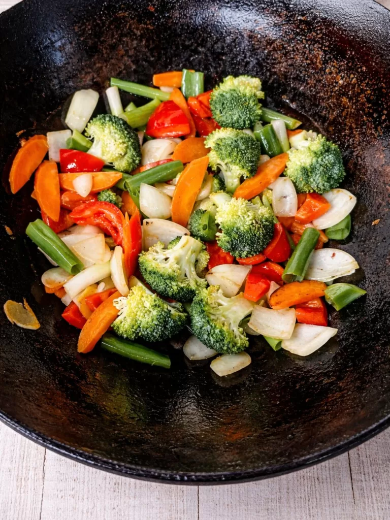 Stir-fried vegetables in a black wok, including broccoli, carrots, red capsicum, onion, and spring onion, cooking over high heat.