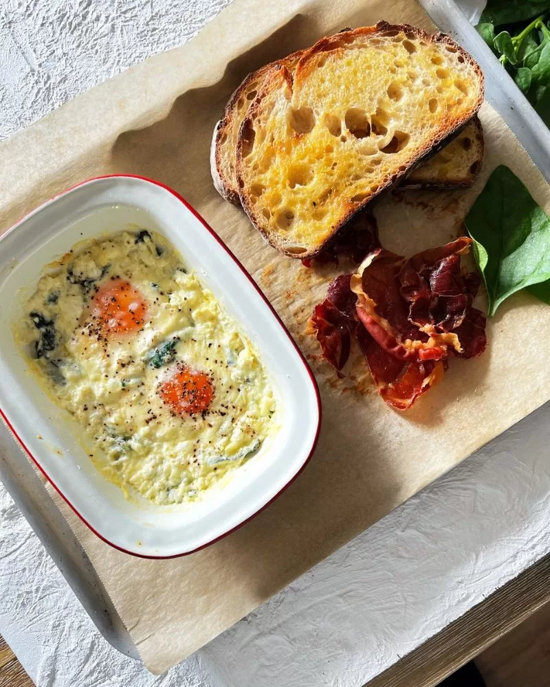 Creamy baked eggs with toast and crispy proscuitto on a lined baking tray.