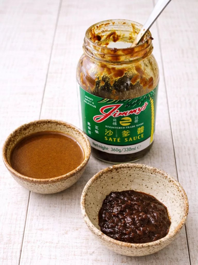 Open jar of Jimmy’s saté sauce on a white timber surface with a spoon inside, served alongside one bowl of smooth saté sauce and one bowl of thick saté paste in natural light.
