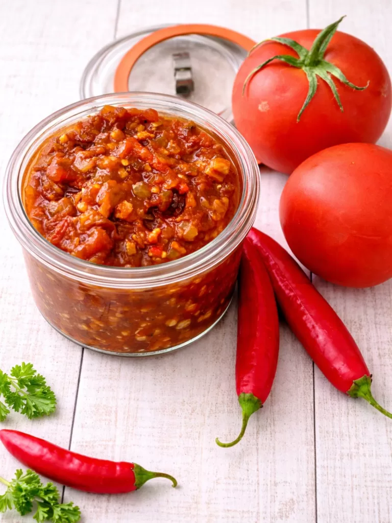 Glass jar of tomato bacon chilli jam on a white timber background, styled with fresh tomatoes and red chillies in soft natural light.