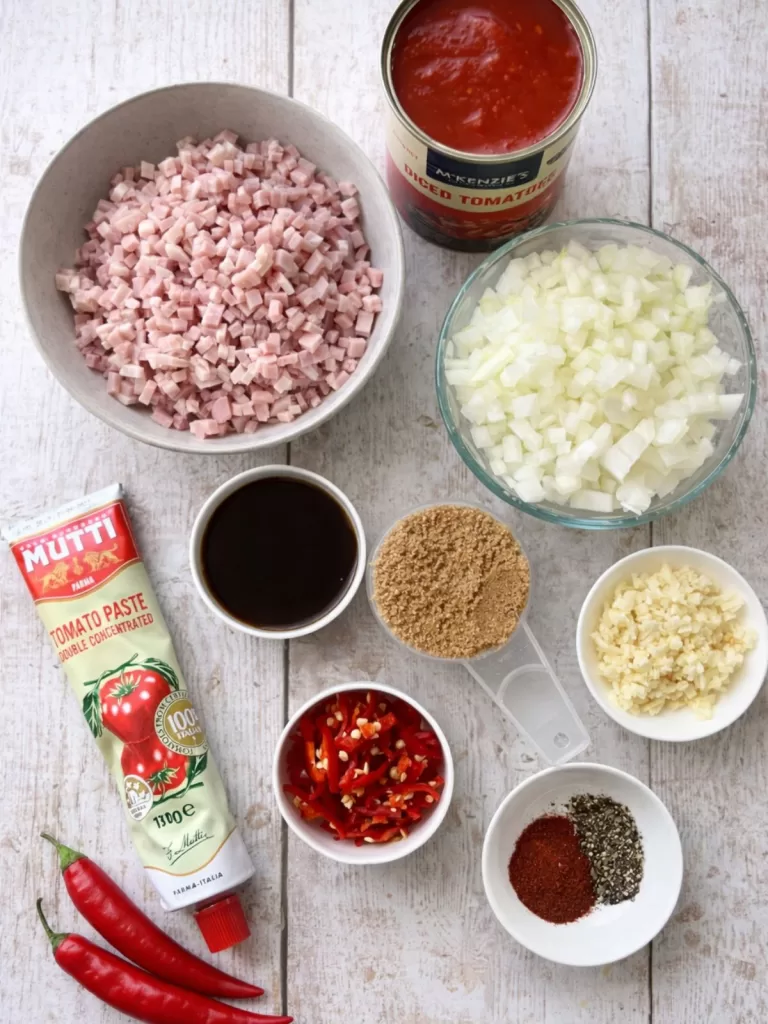 Ingredients for tomato bacon chilli jam arranged on a white timber surface, including diced bacon, chopped onion, canned tomatoes, tomato paste, brown sugar, garlic, chilli and spices in natural light.