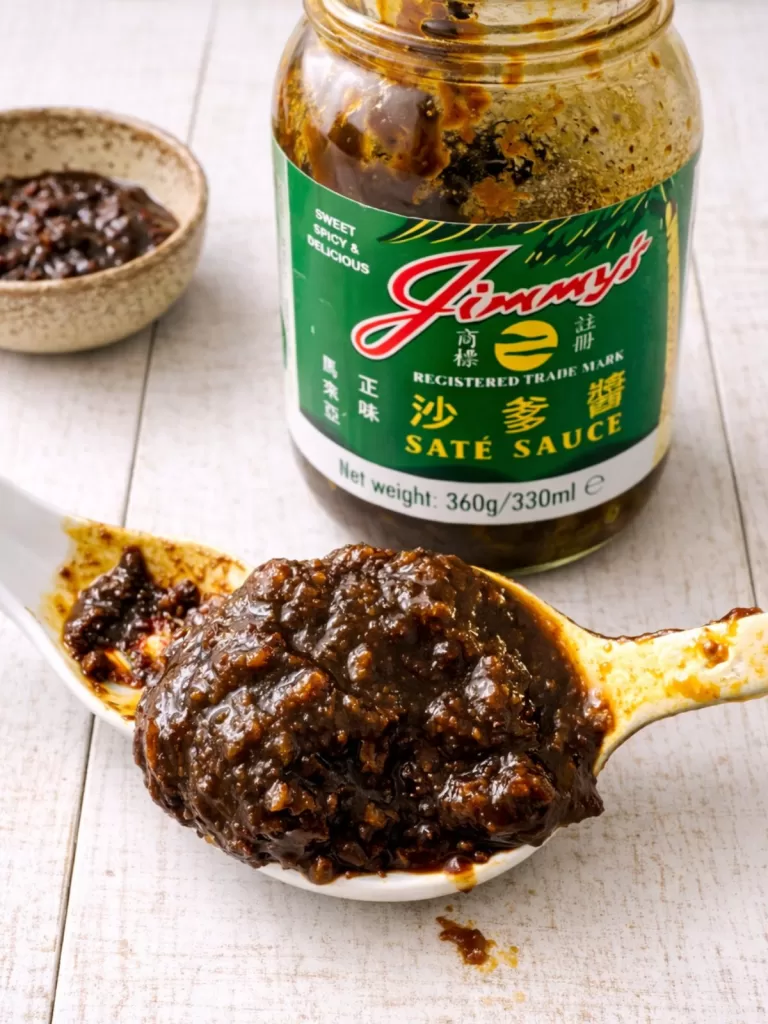 Close-up of a spoonful of thick, chunky satay sauce on a white timber surface, with an open jar of Jimmy’s saté sauce and a small bowl of sauce in the background.