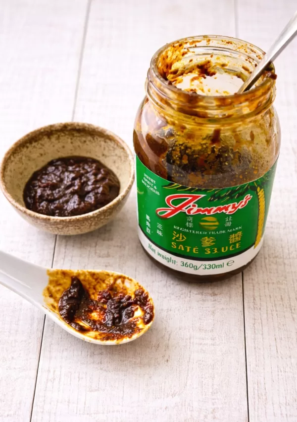 Open jar of Jimmy’s satay sauce on a white timber surface with a small bowl and spoonful of thick, dark satay sauce in natural light.